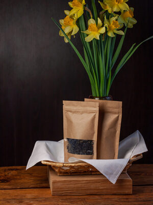 Brown kraft paper pouch bags with coffee beans and tea leaves in wicker basket with white textile on wooden background.